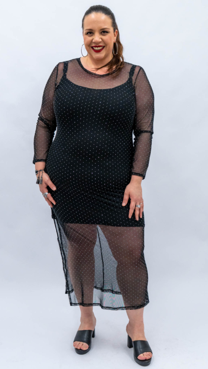 Femme dans une robe noire transparente à pois, debout sur un fond uni, souriante et portant des créoles. Mode Grande taille