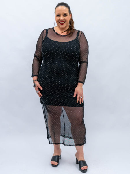 Femme dans une robe noire transparente à pois, debout sur un fond uni, souriante et portant des créoles. Mode Grande taille