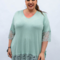 Une femme souriante avec des créoles porte un haut vert menthe avec des manches en dentelle, un pantalon noir, sur un fond uni. Mode Grande taille