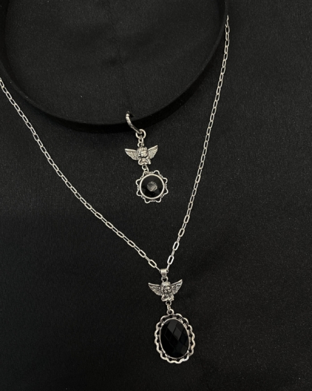 Le « Collier ras de cou anges » présente deux colliers en argent avec des pendentifs sur le thème des anges ; l'un comprend une pierre noire, le tout présenté sur un fond en tissu sombre. Mode Grande taille