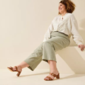 Une femme portant des mules à talons larges marron Gabylou IZA, un chemisier léger et un pantalon vert sauge est assise en souriant sur un fond beige. Mode Grande taille