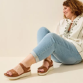 Une femme assise sur le sol sourit, portant un jean bleu clair, un haut blanc brodé et des Sandales pieds larges MIA - Gabylou. Mode Grande taille