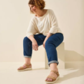 Une femme souriante aux cheveux bouclés porte un haut blanc, un jean et des Sandales pieds larges MIA - Gabylou alors qu'elle est assise sur un bloc sur un fond beige. Mode Grande taille