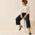 Femme souriante aux cheveux bouclés assise sur un tabouret, portant un haut crème, un pantalon bleu marine et des Sandales à talons pieds larges LEA - Gabylou. Mode Grande taille
