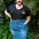 Personne souriante, portant une chemise noire et une jupe en jean, debout à l'extérieur avec de la verdure en arrière-plan. Mode Grande taille