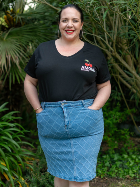 Personne portant une chemise noire « Amour » et une jupe bleue souriante, debout à l'extérieur avec de la verdure en arrière-plan. Mode Grande taille