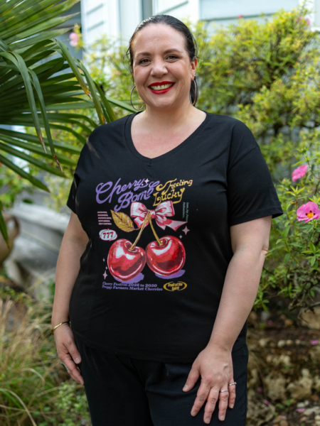 Une personne souriante dans un jardin, portant un t-shirt noir « Cherry Bomb » orné d'un motif de cerises. Fleurs et verdure en arrière-plan. Mode Grande taille