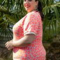 Une femme souriante vêtue d'une chemise à fleurs rose se tient devant des feuilles de palmier, légèrement tournée vers le côté. Mode Grande taille