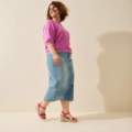 Une femme en haut rose, jupe midi en jean et sandales compensées pieds larges de Gabylou Vic marche sur un fond clair en souriant. Mode Grande taille