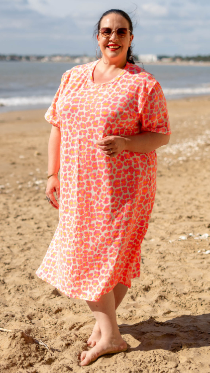 Une femme vêtue d'une robe à motifs rose et orange sourit debout pieds nus sur une plage de sable. Mode Grande taille