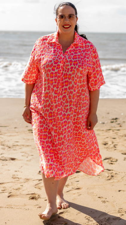 Une femme vêtue d'une robe à motifs rose et orange sourit en marchant pieds nus sur une plage de sable au bord de la mer. Mode Grande taille