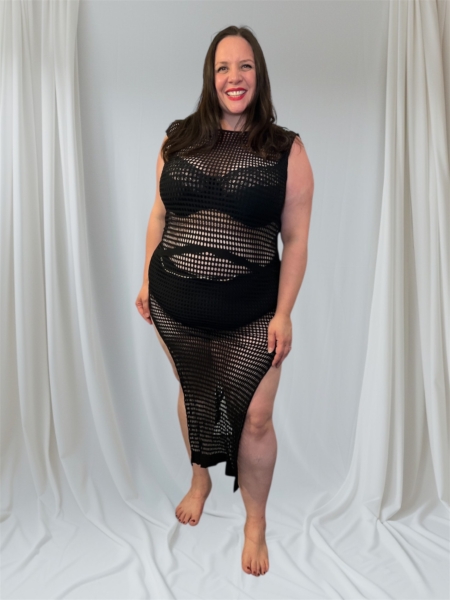 Une femme souriante se tient pieds nus devant des rideaux drapés blancs, portant la Robe résille fendue sur les côtés en noir. Mode Grande taille