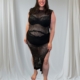 Une femme souriante se tient pieds nus devant des rideaux drapés blancs, portant la Robe résille fendue sur les côtés en noir. Mode Grande taille