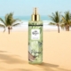 Une bouteille de Brume Pistache Body Splash repose sur une plage de sable, avec des palmiers et l'océan en arrière-plan. Mode Grande taille