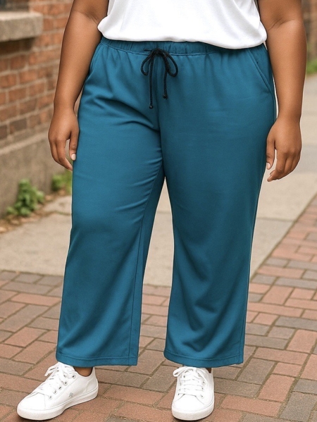 Une personne portant le Pantalon décontracté en jersey de couleur sarcelle, associé à des baskets blanches et à un haut blanc, se tient sur un trottoir en briques. Mode Grande taille