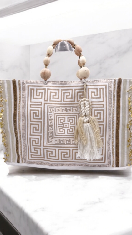 Sac à main beige et or avec pompon, anse en perles de bois et motif de clé grecque, présenté sur une surface en marbre. Mode Grande taille