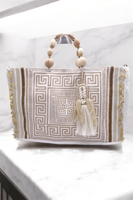 Sac à main beige et or avec pompon, anse en perles de bois et motif de clé grecque, présenté sur une surface en marbre. Mode Grande taille