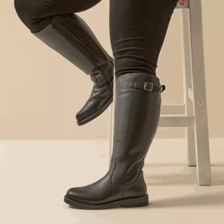 Personne portant Gabylou Bottes cavalières mollets larges 4XL Kate en noir avec un pantalon foncé, assise sur un tabouret blanc sur fond beige. Mode Grande taille