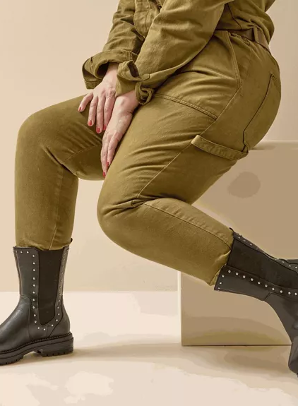 Personne en combinaison olive assise, présentant les Bottines à clous pour pieds larges 2XL Jade by Gabylou - bottes noires cloutées à semelles épaisses - sur un fond beige. Mode Grande taille