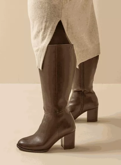Une personne portant les Bottes à talon mollets larges 4/5XL Gladys - Gabylou, des bottes brunes à talon au niveau du genou, associées à une jupe beige avec une fente sur le devant. Mode Grande taille