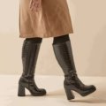 Une personne portant des Bottes mollets larges 2XL Mélissa - Gabylou, des bottes noires texturées au genou et un manteau beige marche à l'intérieur. Mode Grande taille