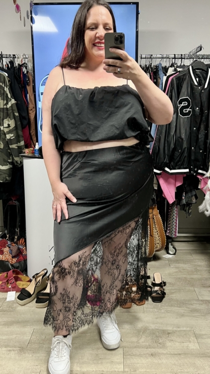 Une femme souriante prend un selfie dans un miroir, vêtue d'un crop top noir et d'une Jupe satin dentelle noire, entourée de vêtements et d'accessoires. Mode Grande taille