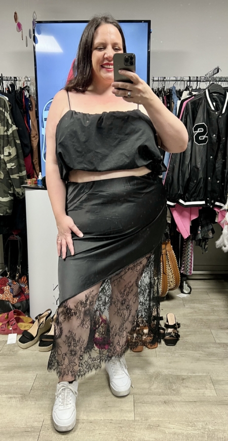 Une femme souriante prend un selfie dans un miroir, vêtue d'un crop top noir et d'une Jupe satin dentelle noire, entourée de vêtements et d'accessoires. Mode Grande taille