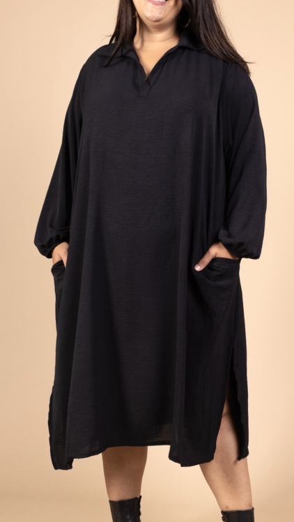 Une femme portant la Robe col chemise fendue sur les côtés, une robe noire ample avec des manches longues et des poches, se tient sur un fond beige. Mode Grande taille