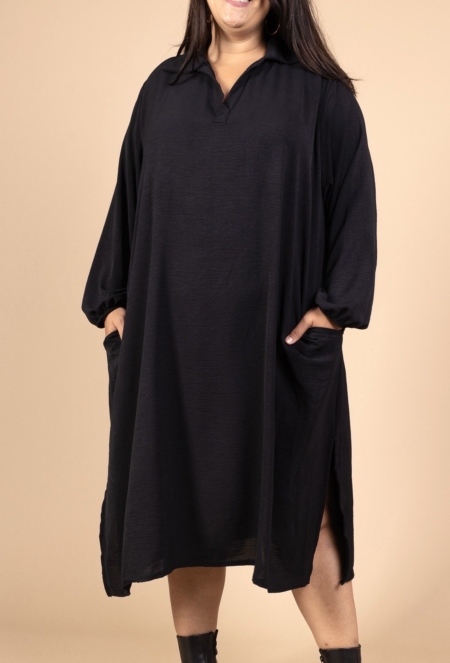Une femme portant la Robe col chemise fendue sur les côtés, une robe noire ample avec des manches longues et des poches, se tient sur un fond beige. Mode Grande taille