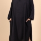 Une femme portant la Robe col chemise fendue sur les côtés, une robe noire ample avec des manches longues et des poches, se tient sur un fond beige. Mode Grande taille