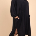 Une femme souriante portant la "Robe col chemise fendue sur les côtés", une robe noire ample à manches longues avec des poches, associée à des bottines noires, pose sur un fond beige. Mode Grande taille