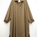 Une robe beige, ample et à manches longues, appelée "Robe col chemise fendue sur les côtés", avec un col, est suspendue à un cintre noir contre un mur blanc. Mode Grande taille