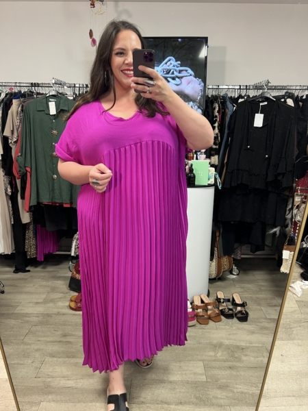 Une femme sourit dans un magasin de vêtements, portant la Robe longue plissée violine - une robe plissée violet vif - entourée de vêtements et de chaussures suspendus. Mode Grande taille