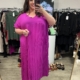 Une femme sourit dans un magasin de vêtements, portant la Robe longue plissée violine - une robe plissée violet vif - entourée de vêtements et de chaussures suspendus. Mode Grande taille