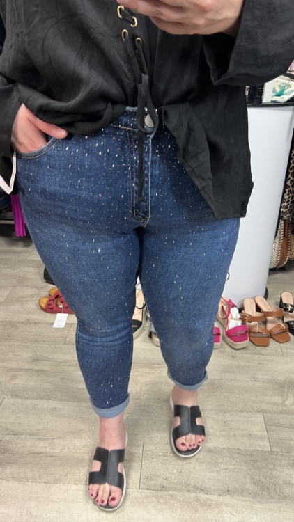 Une personne porte la Jean à strass, associée à un haut noir et à des sandales, debout dans un magasin de vêtements avec des chaussures exposées en arrière-plan. Mode Grande taille